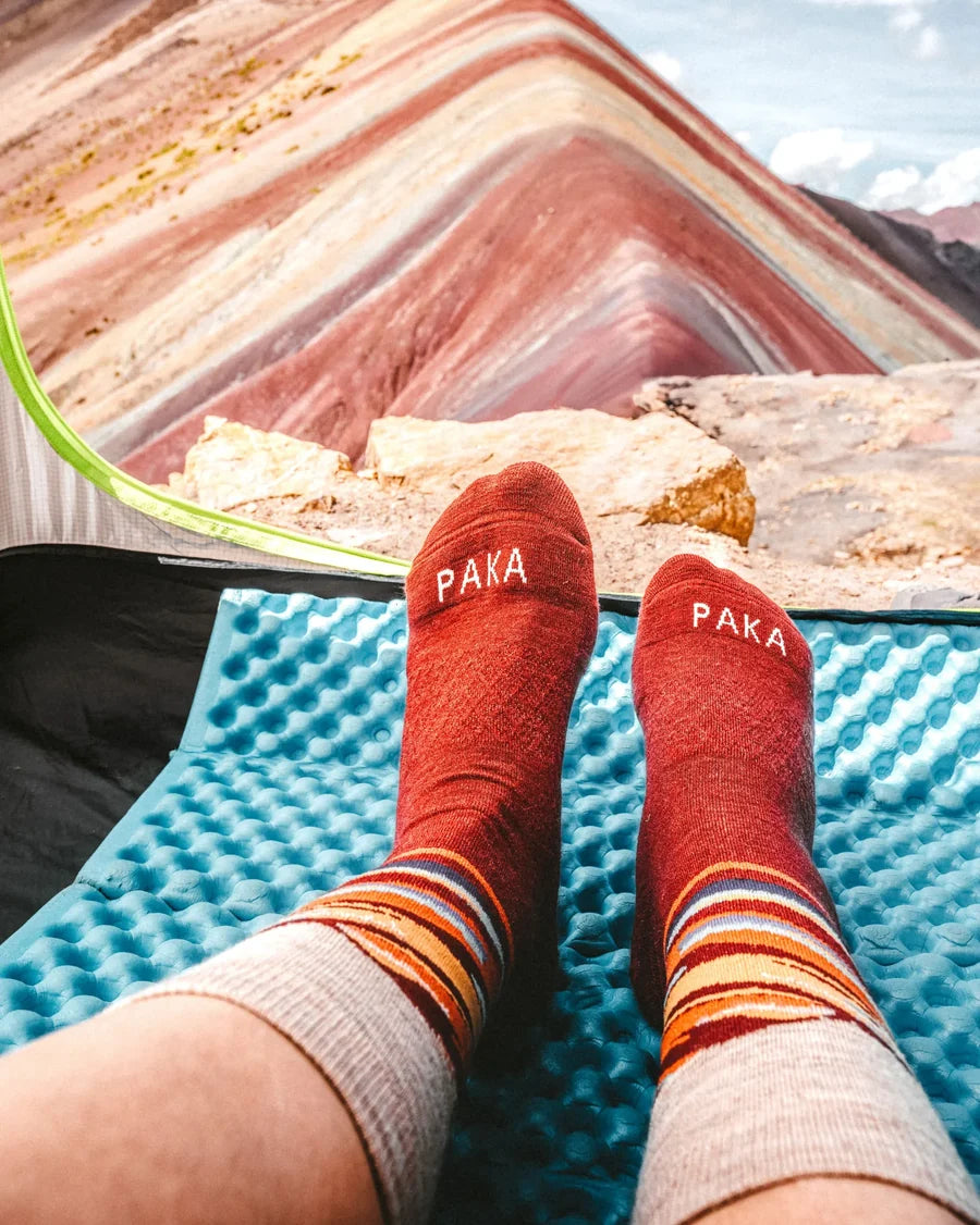 A person's legs resting on a blue sleeping pad with a colorful striped mountain landscape in the background. The person is wearing red crew socks with 'PAKA' embroidered on the toes and striped cuffs.