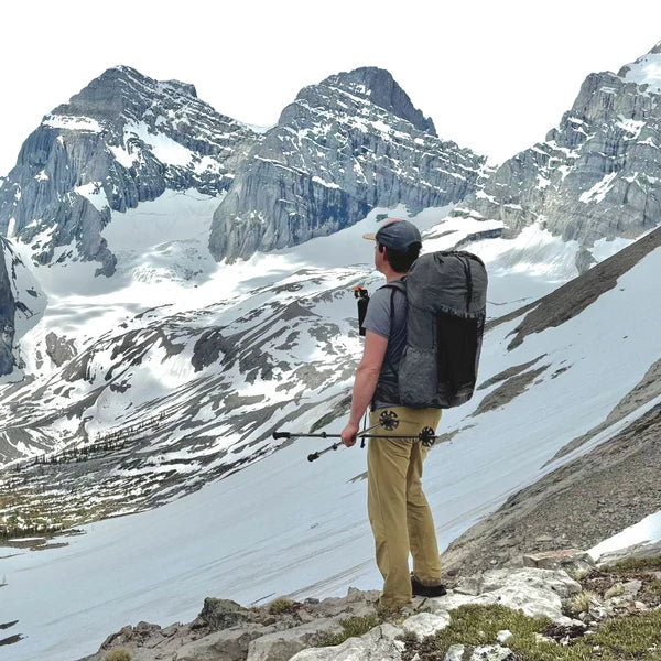 A hiker with a large black backpack and trekking poles stands on a rocky mountain ridge overlooking a vast snow-covered alpine valley and jagged peaks.