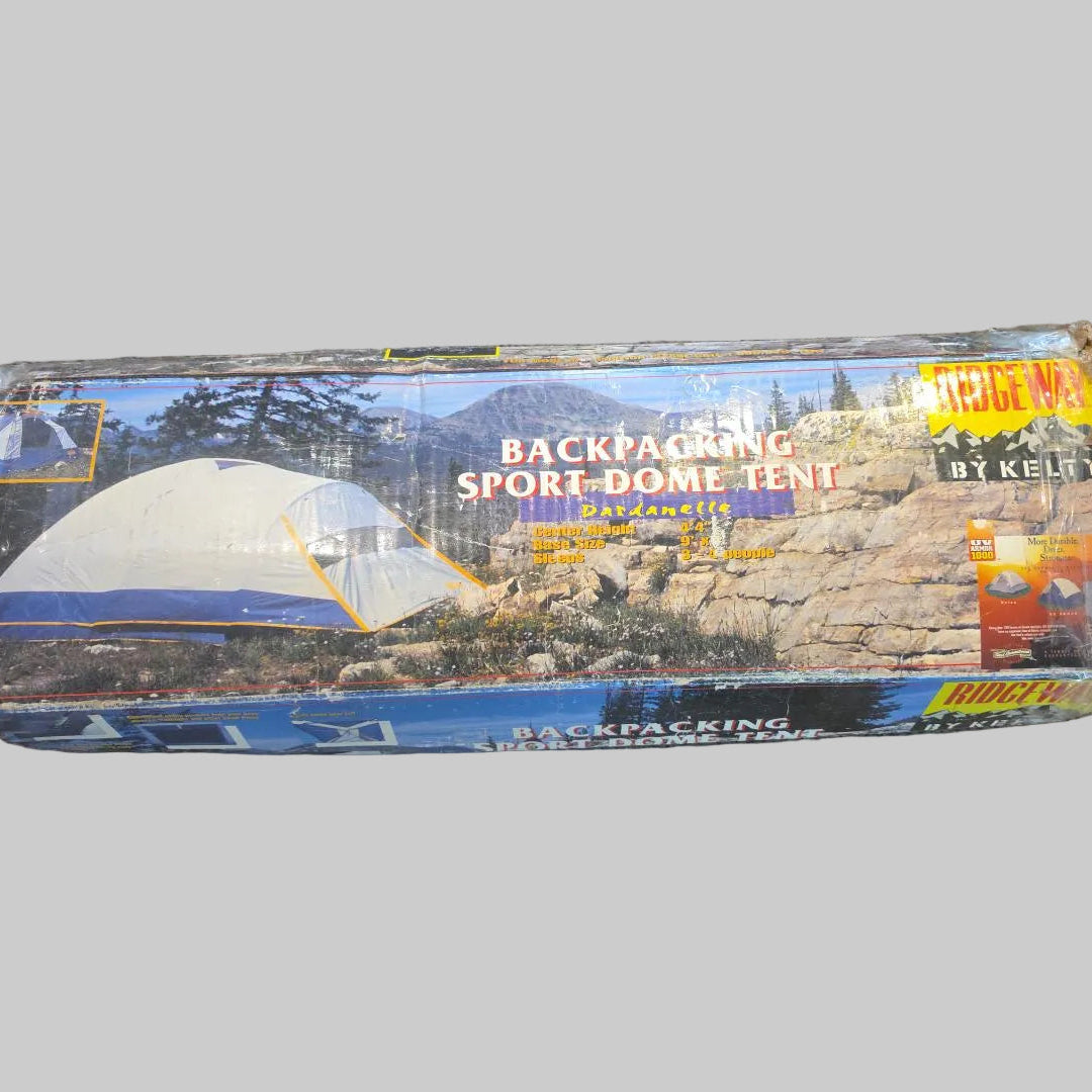 A packaged backpacking sport-dome tent box by Kelly, wrapped in clear plastic, lying on a brown carpeted floor. The box features a graphic of a white and blue dome tent in a mountain setting.