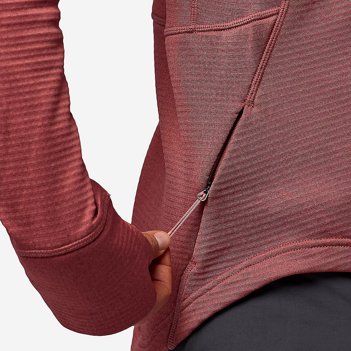 Close-up of a person wearing a ribbed dusty rose zippered hoodie, showing the sleeve, pocket, and hand pulling the zipper, paired with dark gray pants against a light gray background.
