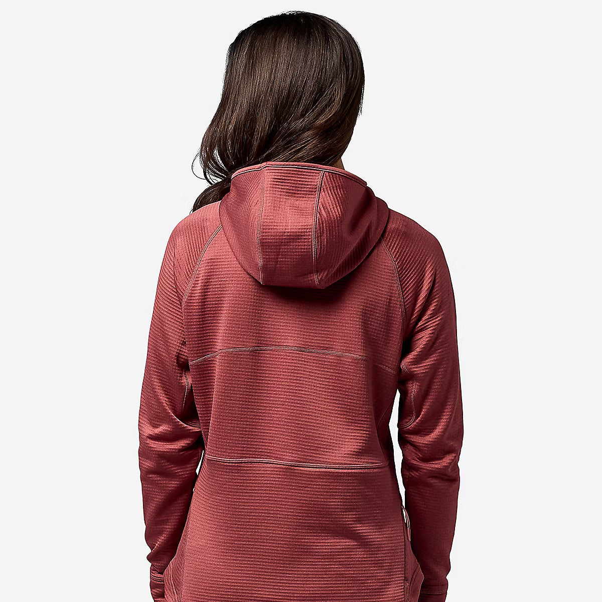 A woman viewed from the back wearing a textured, ribbed hooded athletic jacket in a muted brick red color, paired with dark gray pants, against a plain light gray background.