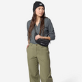 A woman modeling olive green wide-leg pants, a dark gray long-sleeve henley shirt, a black beanie, and a black crossbody fanny pack against a light gray studio background.