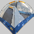 A dome-style camping tent pitched on grass outdoors, with tree leaves casting shadows across the fabric. The tent is primarily light gray with dark blue lower panels and orange trim accents.