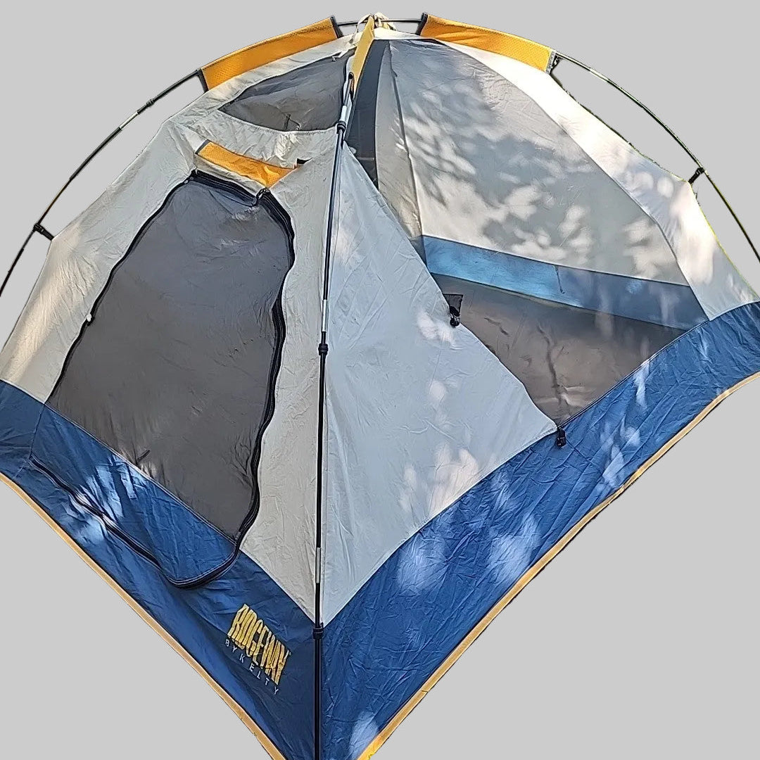 A dome-style camping tent pitched on grass outdoors, with tree leaves casting shadows across the fabric. The tent is primarily light gray with dark blue lower panels and orange trim accents.