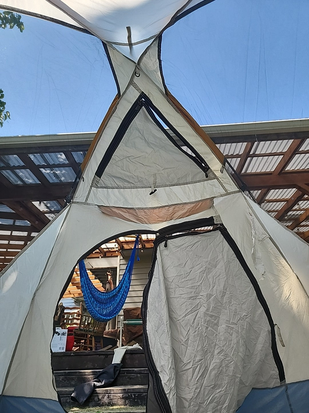 A large white and gray dome-style camping tent with black trim, shown open with a blue hammock hanging inside. The tent is photographed outdoors under a clear blue sky, with a wooden roof structure and patio furniture visible in the background.